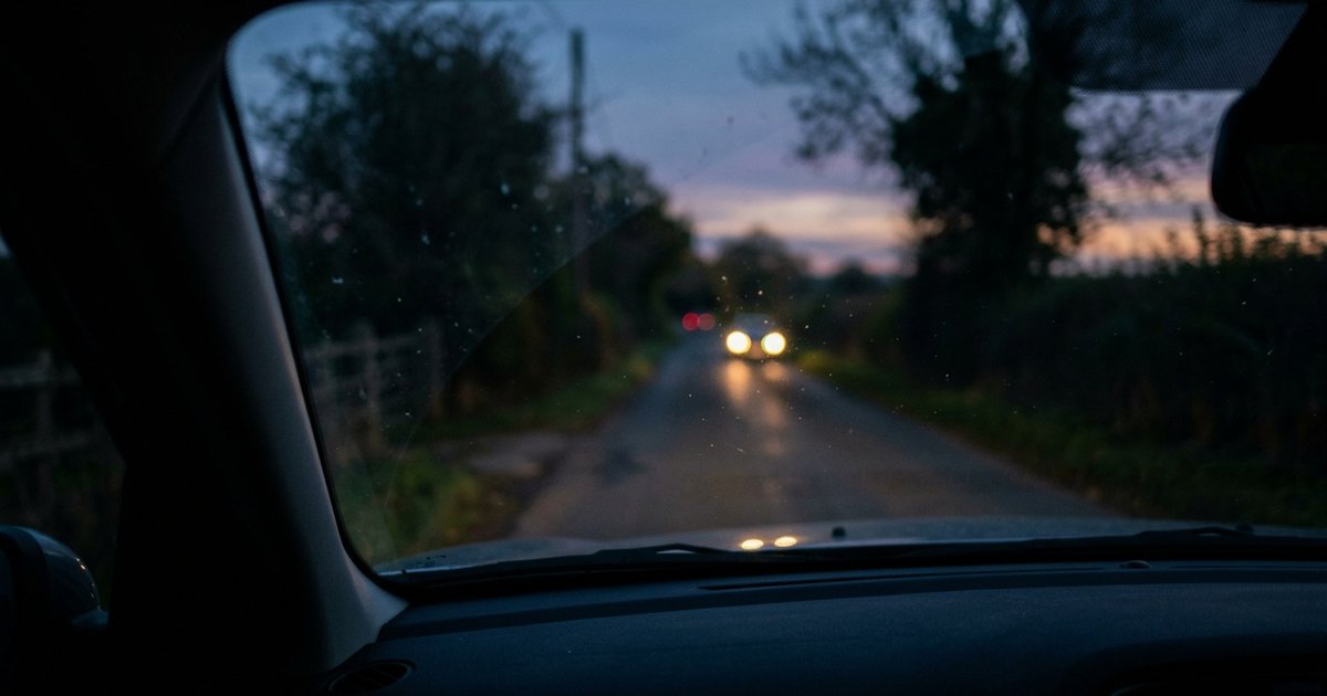 A quiet country road at dusk seen through a car windscreen, soft blurred oncoming headlights, low-light evening atmosphere